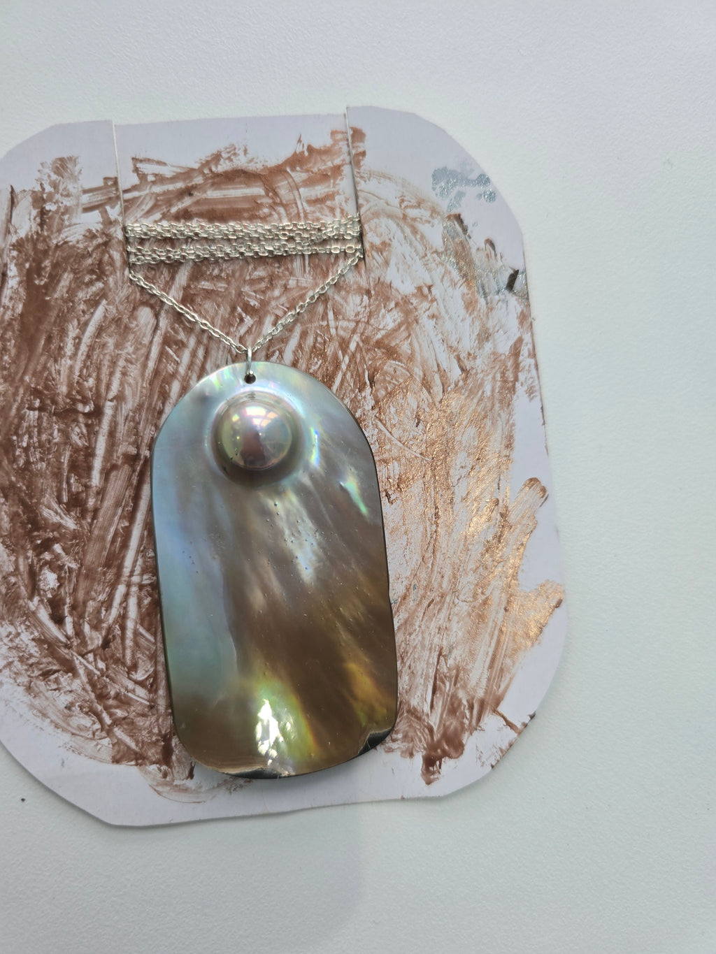A necklace with a large iridescent mother-of-pearl pendant and a silver-toned chain, displayed on a white card with brown paint smears against a light gray wall.