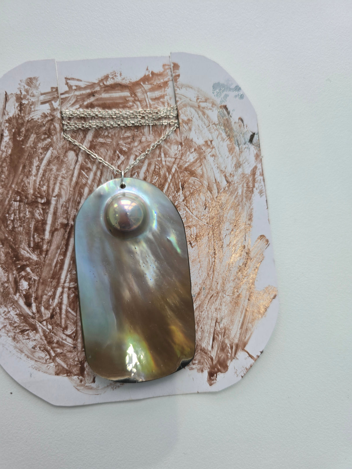 A necklace with a large iridescent mother-of-pearl pendant and a silver-toned chain, displayed on a white card with brown paint smears against a light gray wall.